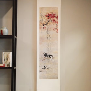 May include: A vertical wall hanging scroll featuring a painting of a bird swimming in water beneath a tree with red blossoms. The artwork is rendered in a soft, muted color palette on a light background, suspended from a metal hook at the top.