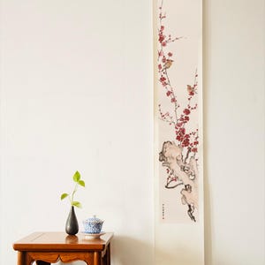 May include: A vertical wall scroll featuring a painting of a gnarled tree branch with red blossoms and two small birds. Beside it, a wooden side table holds a small black vase with a green plant and a blue and white patterned lidded tea bowl.