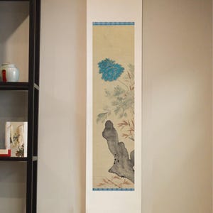 May include: A vertical wall hanging scroll featuring a painting of a bright blue chrysanthemum flower, green foliage, and a dark grey rock formation. The artwork is mounted on a white paper backing with blue patterned borders at the top and bottom.