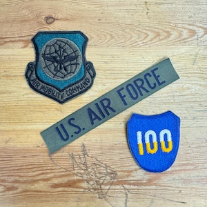 Peut inclure: Trois patchs militaires sur une surface en bois. Un patch bleu et vert avec un globe et le texte "Air Mobility Command". Un patch vert avec le texte "U.S. Air Force". Un patch bleu avec un contour blanc et le numéro "100" en jaune.