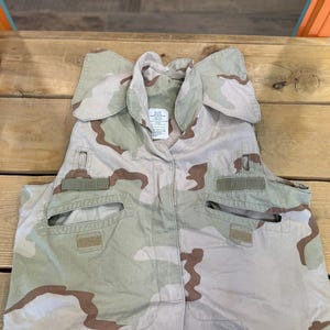 Peut inclure: Un gilet de camouflage beige, marron et vert avec plusieurs poches et une fermeture à bouton. Le gilet porte une étiquette indiquant "Made in USA".