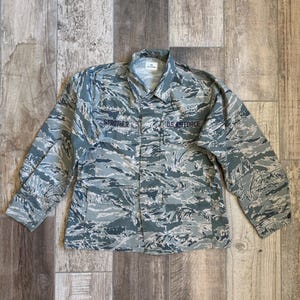 Peut inclure: Veste d'uniforme de l'armée de l'air à manches longues et boutonnée, dans un motif de camouflage bleu et gris. Le nom "STROTHER" est brodé au-dessus de la poche de poitrine gauche, et "U.S. AIR FORCE" au-dessus de la poche droite.
