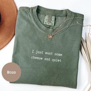 Op de afbeelding: Een mosgroen t-shirt met de tekst "I just want some cheese and quiet" in het wit. Een gouden ketting met een kleine hanger ligt op het shirt. Een bruine hoed en een gedroogde plant staan op de achtergrond.