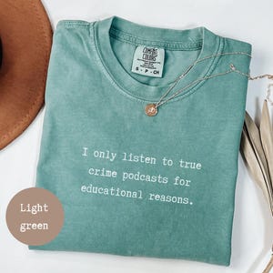 Può includere: T-shirt verde chiaro con la scritta "I only listen to true crime podcasts for educational reasons." Una collana d'argento con un ciondolo è drappeggiata sulla maglietta. La maglietta è piegata su una superficie bianca.