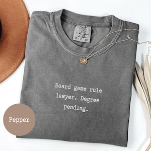 May include: A grey Comfort Colors t-shirt with the text "Board game rule lawyer. Degree pending." A gold necklace with a pendant is on the shirt. A brown hat is in the upper left corner. The shirt is labelled "Pepper."