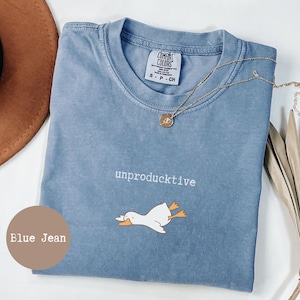 May include: A blue jean coloured t-shirt with a crew neckline and a Comfort Colors brand label. The shirt features the white text "unproductive" above a graphic of a white duck with orange feet and a beak, lying flat on its stomach.