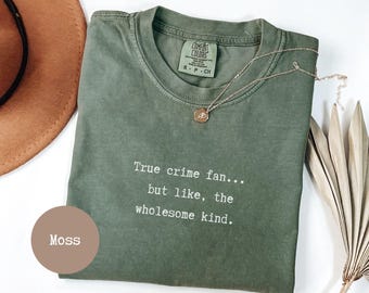 Op de afbeelding: Een mosgroen t-shirt met de tekst "True crime fan... but like, the wholesome kind." Een gouden ketting met een hanger ligt op het shirt. Een bruine hoed en een gedroogde plant staan op de achtergrond.