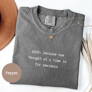 Op de afbeelding: Een grijs Comfort Colors T-shirt met de tekst "ADHD: because one thought at a time is for amateurs" in het wit. Een gouden ketting met een hanger ligt op het shirt. Een bruine hoed is zichtbaar op de achtergrond.
