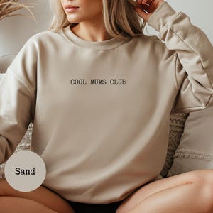 Pode incluir: Um moletom cor de areia com o texto "COOL MUMS CLUB" em preto. O moletom tem gola redonda e mangas compridas. A imagem também inclui um rótulo redondo que diz "Sand".