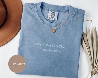 Op de afbeelding: Een blauw spijkerbroekkleurig t-shirt met de tekst "TRUE CRIME DETECTIVE COUCH DIVISION" in het wit. Een gouden ketting met een hanger is over het shirt gedrapeerd. Het shirt is opgevouwen en heeft een Comfort Colors-label.
