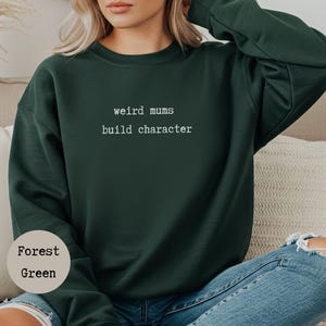 Op de afbeelding: Boszwarte sweatshirt met de witte tekst "weird mums build character". De sweatshirt heeft een ronde hals en is effen van kleur. Het model draagt een blauwe jeans.