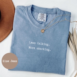 Könnte beinhalten: Ein blaues Jeans-T-Shirt mit dem weißen Aufdruck "Less talking. More snacking.". Eine goldene Halskette mit Anhänger liegt auf dem Shirt. Das Shirt ist gefaltet und hat ein Comfort Colors-Etikett.