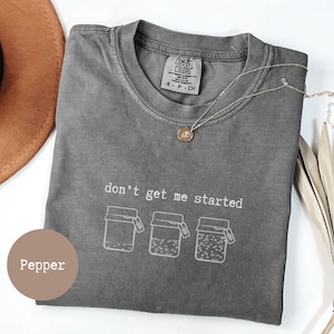 May include: A charcoal gray short-sleeve t-shirt with a crew neckline. The shirt features white line art of three jars with tags and the text "don't get me started" printed above them. The interior neck label reads "Comfort Colors" and "S-P-CH".