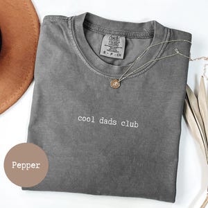 May include: A grey Comfort Colors t-shirt with the text "cool dads club" in white. A gold necklace with a pendant rests on the shirt. A brown hat is visible in the upper left corner.