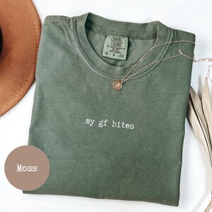 May include: A moss green t-shirt with the text "my gf bites" in white. A gold necklace with a small pendant rests on the shirt. A brown hat and dried palm leaves are in the background.