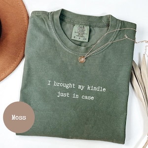 Pode incluir: Uma camiseta verde musgo com o texto branco "I brought my kindle just in case". A camisa tem gola redonda e uma pequena etiqueta na gola. Um colar de ouro com um pingente está sobre a camisa.