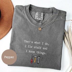 Puede incluir: Una camiseta de manga corta de color gris carbón con el texto en blanco "That's what I do. I fix stuff and I know things." y un gráfico de un destornillador rojo, un martillo amarillo y una llave inglesa gris. La camiseta tiene una etiqueta Comfort Colors.