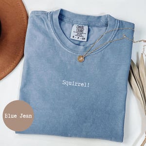 Puede incluir: Una camiseta azul vaquero con la palabra "Squirrel!" bordada en blanco. La camiseta tiene cuello redondo y una pequeña etiqueta en el cuello. Un collar dorado con un colgante está drapeado sobre la camiseta.