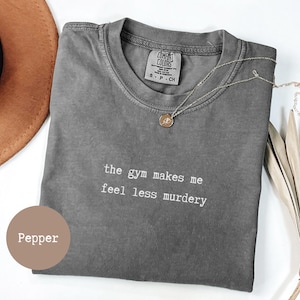 Op de afbeelding: Een grijs Comfort Colors T-shirt met de tekst "the gym makes me feel less murdery" in het wit. Een gouden ketting met een hanger ligt op het shirt. Een bruine hoed staat op de achtergrond.