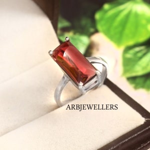 Può includere: Un anello d'argento con una grande gemma rossa rettangolare. L'anello ha una fascia semplice e un'incastonatura che tiene saldamente la gemma. Il testo "ARBJWELLERS" è visibile in basso nell'immagine.