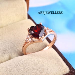 May include: Rose gold ring featuring a heart-shaped, deep red gemstone. The ring is accented with small, clear gemstones on either side of the heart. The ring is displayed in a jewelry box. The text "ARBJEWELLERS" is visible.