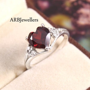 Peut inclure: Une bague en argent avec une pierre rouge en forme de cœur et de plus petites pierres claires. La bague est dans une boîte blanche avec le texte "ARBJewellers".