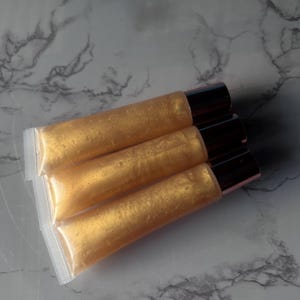 May include: Three tubes of gold lip gloss with rose gold caps. The tubes are stacked on top of each other.