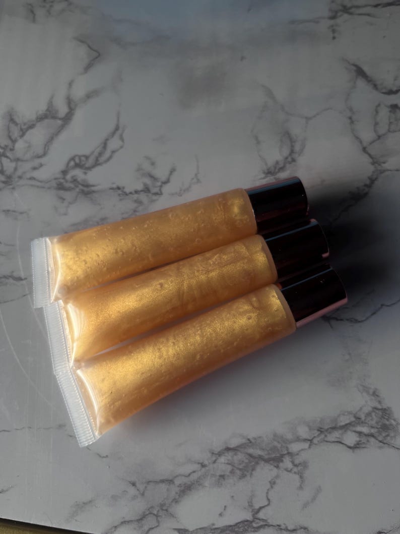 May include: Three tubes of gold lip gloss with rose gold caps. The tubes are stacked on top of each other.