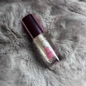 May include: A clear glass bottle with a pink and silver cap filled with a clear liquid with pink and silver glitter. The bottle is sitting on a gray fuzzy surface.