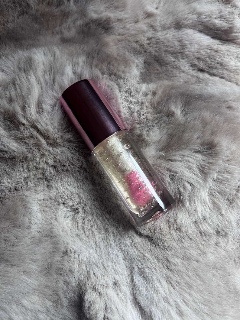 May include: A clear glass bottle with a pink and silver cap filled with a clear liquid with pink and silver glitter. The bottle is sitting on a gray fuzzy surface.