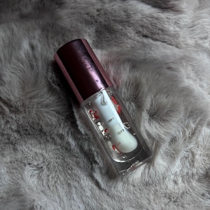 May include: A clear lip gloss tube with a rose gold cap. The gloss contains red glitter and is partially filled.