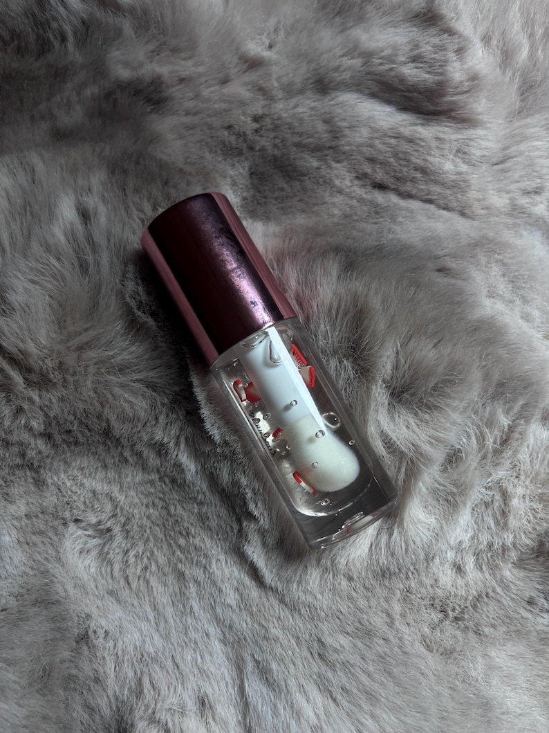 May include: A clear lip gloss tube with a rose gold cap. The gloss contains red glitter and is partially filled.