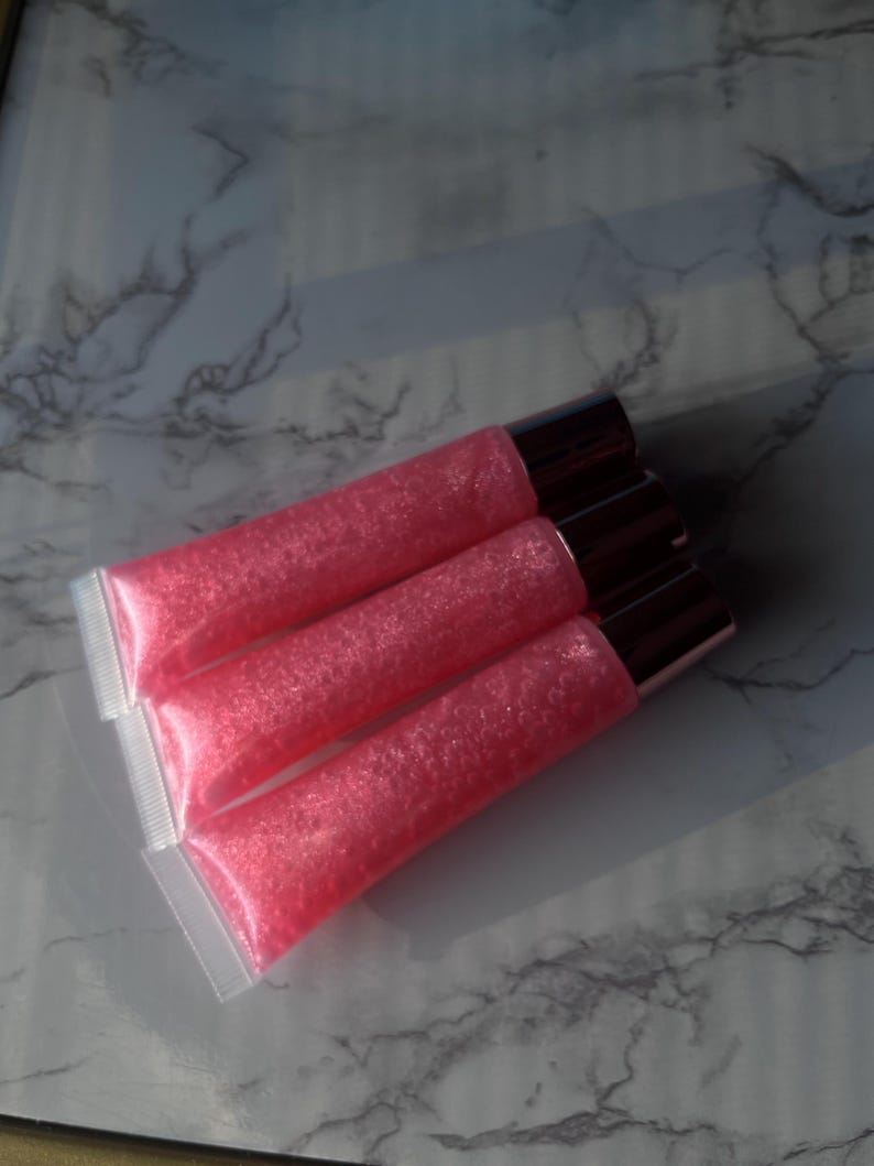 May include: Three pink lip gloss tubes with a shimmery finish. The tubes are clear plastic with a rose gold cap.