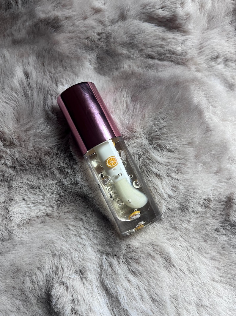 May include: A clear glass bottle with a pink lid containing a clear liquid with a small yellow citrus fruit inside. The bottle is sitting on a gray fuzzy surface.