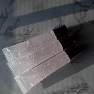 May include: Three clear lip gloss tubes with silver caps. The tubes are stacked on top of each other and are filled with a clear, slightly pink liquid.