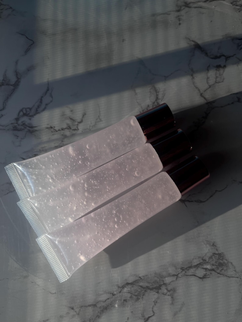 May include: Three clear lip gloss tubes with silver caps. The tubes are stacked on top of each other and are filled with a clear, slightly pink liquid.