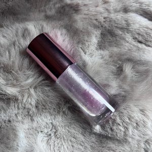 May include: A clear glass bottle with a pink and silver iridescent liquid inside. The bottle has a pink cap and is sitting on a gray fuzzy surface.