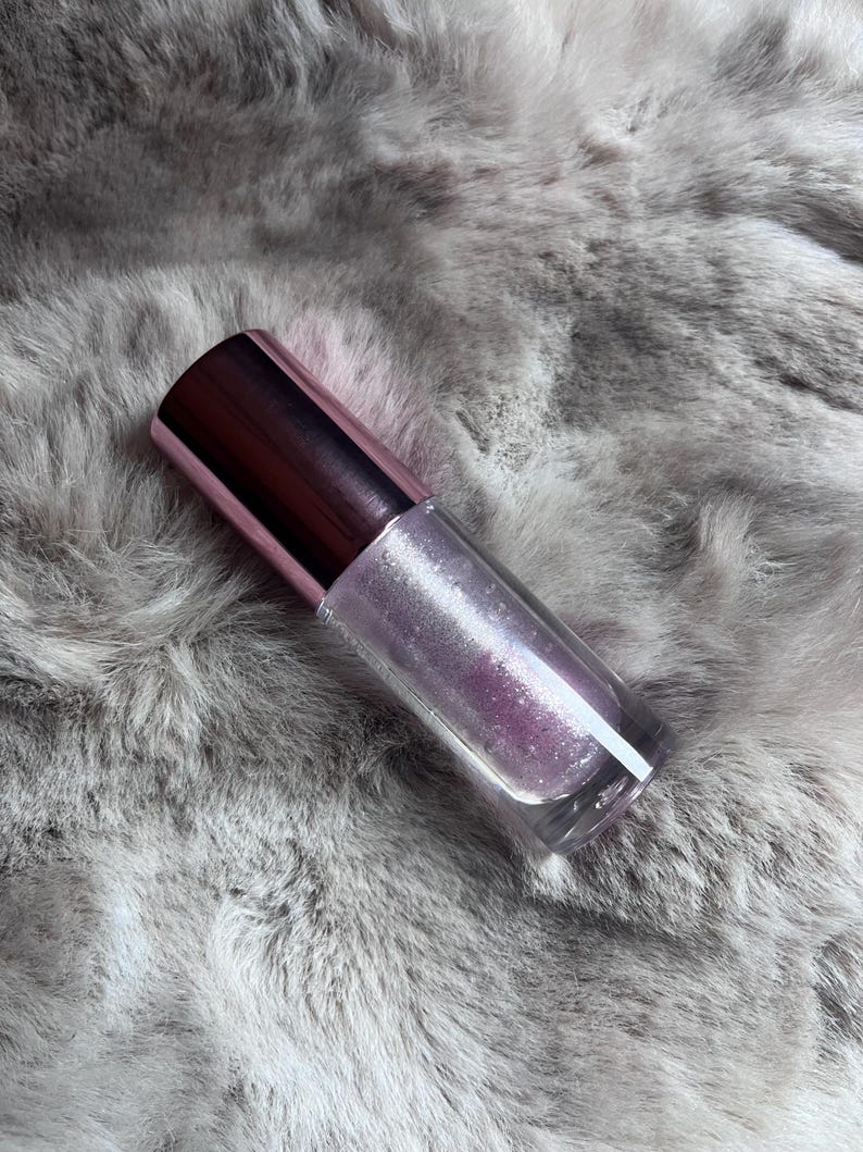 May include: A clear glass bottle with a pink and silver iridescent liquid inside. The bottle has a pink cap and is sitting on a gray fuzzy surface.