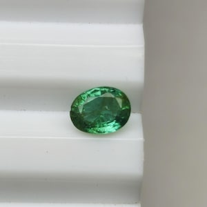 May include: A vivid green, oval-shaped gemstone. The gem is displayed on a white, ridged surface. The facets of the stone reflect light, creating a sparkling effect. This gemstone is suitable for jewellery.