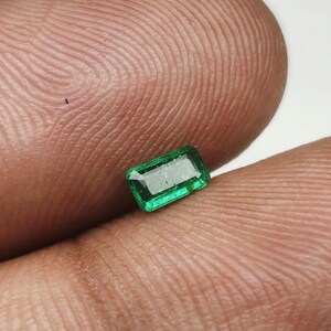 Può includere: Una piccola gemma di smeraldo rettangolare con una vivace tonalità verde. Lo smeraldo è tenuto tra due dita, mostrando il suo taglio e la sua purezza. Le sfaccettature della gemma riflettono la luce, evidenziandone la brillantezza. Lo sfondo è di un colore neutro, bianco sporco.