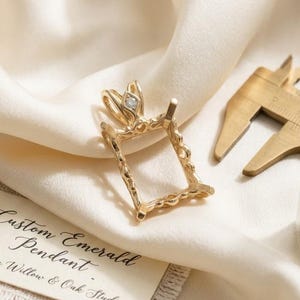 May include: A gold pendant setting with a rectangular frame and a small diamond accent at the top. A tag reads Custom Emerald Pendant The Willow & Oak Studio. A brass caliper and a small glass vial containing green moss are also visible.
