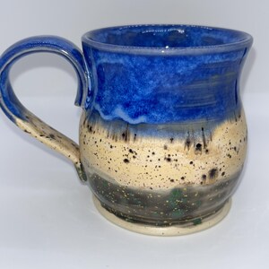 May include: A handmade ceramic mug with a blue, tan, and black speckled design. The top of the mug is a deep blue, transitioning to tan with black speckles, and a dark base. The mug has a curved handle.