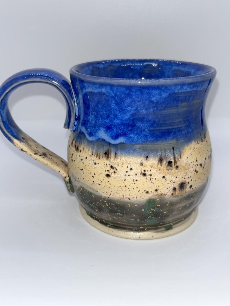 May include: A handmade ceramic mug with a blue, tan, and black speckled design. The top of the mug is a deep blue, transitioning to tan with black speckles, and a dark base. The mug has a curved handle.