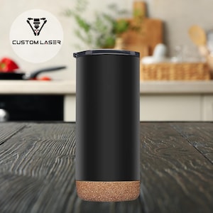 May include: A black tumbler with a cork base and a black lid. The tumbler has the text "CUSTOM LASER" and a logo in a white circle. The tumbler is cylindrical and sits on a dark wooden surface.