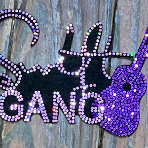 May include: A black cut-out design with the words "Soulful Gang" and a purple rhinestone guitar. The letters and guitar are outlined with purple rhinestones. The background is a weathered wood texture.