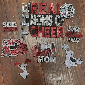 May include: A selection of rhinestone-embellished cheerleading-themed designs. The designs feature phrases such as "REAL MOMS OF CHEER", "SEE RED", and "BLACK GIRL CHEER". Cheerleader figures and pom-pom graphics are also present.