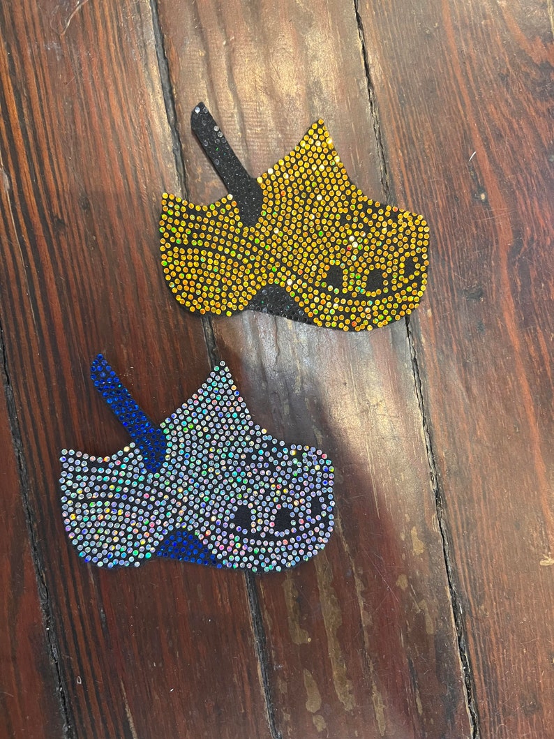May include: Two decorative clog-shaped shoe charms, one yellow and one blue, both embellished with sparkling rhinestones. The charms have black straps and are set against a dark wood grain background.