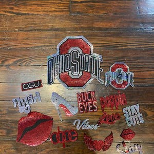 Puede incluir: Una colección de parches y apliques con incrustaciones de pedrería, que incluyen logotipos de "Ohio State", labios, un tacón alto y frases como "Buck Eyes" y "Stay Cute". Los artículos son rojos, plateados y negros, sobre una superficie de madera.