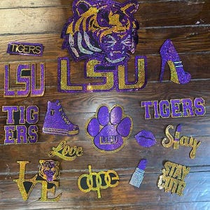 Può includere: Una collezione di oggetti ricoperti di strass, tra cui una testa di tigre, le lettere "LSU" e "TIGERS", una scarpa con tacco alto, un'impronta di zampa e le parole "Love", "Dope", "Stay" e "Stay Cute". Gli oggetti sono viola, oro e argento.