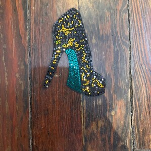 May include: A high-heeled shoe-shaped decorative item. The shoe is black, covered in small, sparkling embellishments. The heel and toe area are teal, with yellow and silver accents throughout the design. The item is set against a wooden background.